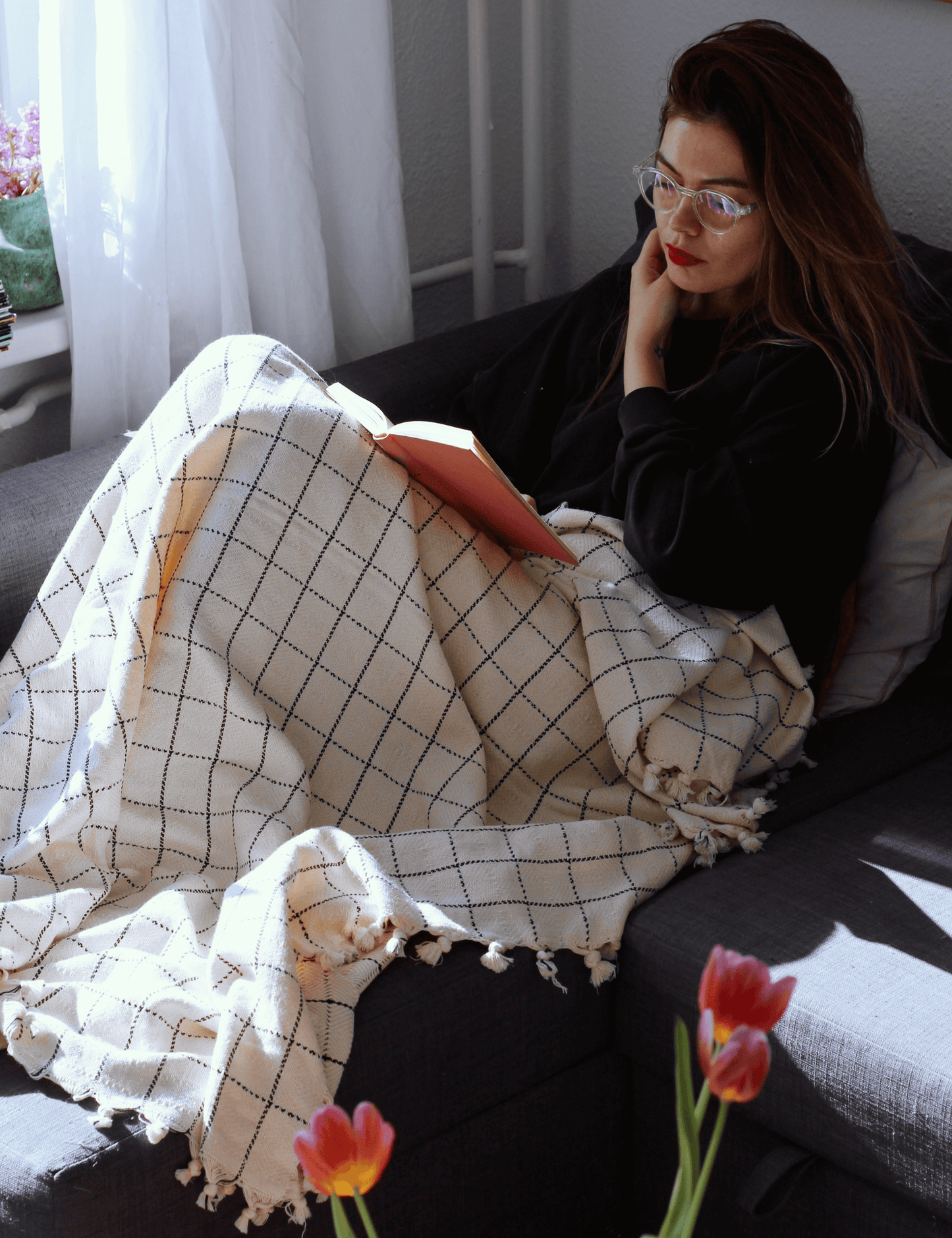 Woman reading with handwoven cotton throw blanket in windowpane pattern, adding coziness to home decor.